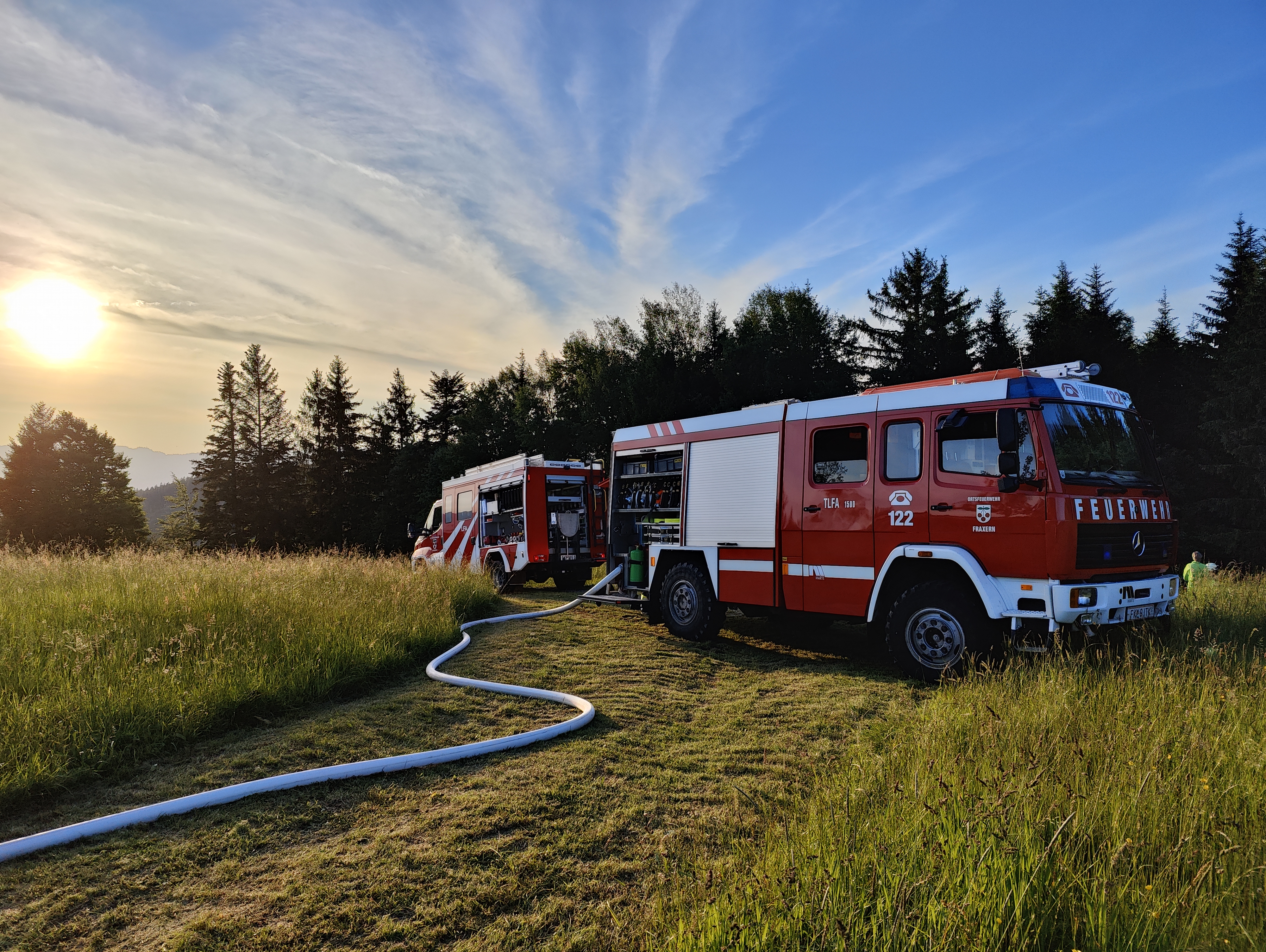 TLFA 1500 und Löschfahrzeug der Ortsfeuerwehr Fraxern bei Sonnenuntergang, Schlauchleitung über die Wiese